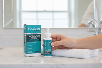 洗面台の上の箱と折り畳んだタオルの横で、FOLIGAIN DHTセラムを持つ手。