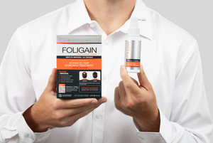 ミノキシジル5%とTRIOXIDIL 5%配合のFOLIGAIN外用トリートメントの箱とスプレーボトルを持つ男性。